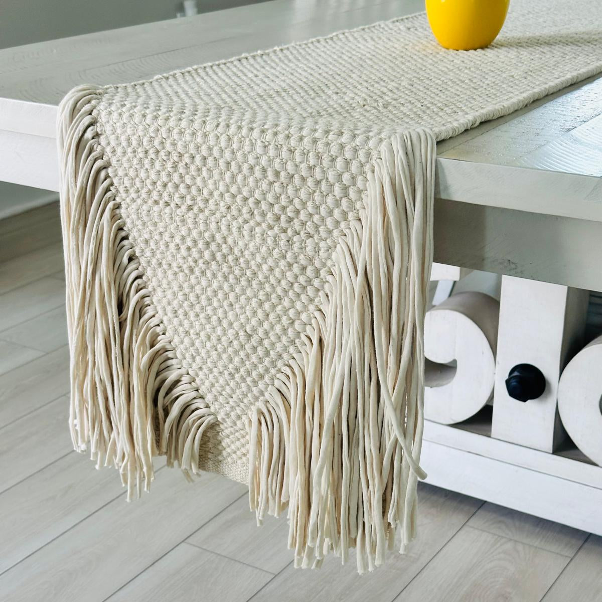 Beige woven table runner with fringe on a wooden table.