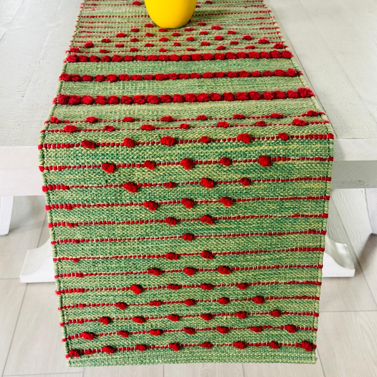 Green and red woven table runner on a white surface with a yellow vase.
